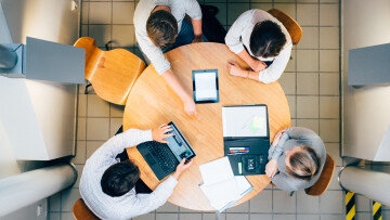 Bild tagen ovanifrån, 4 personer sitter runt ett bor med dator och anteckningsmaterial